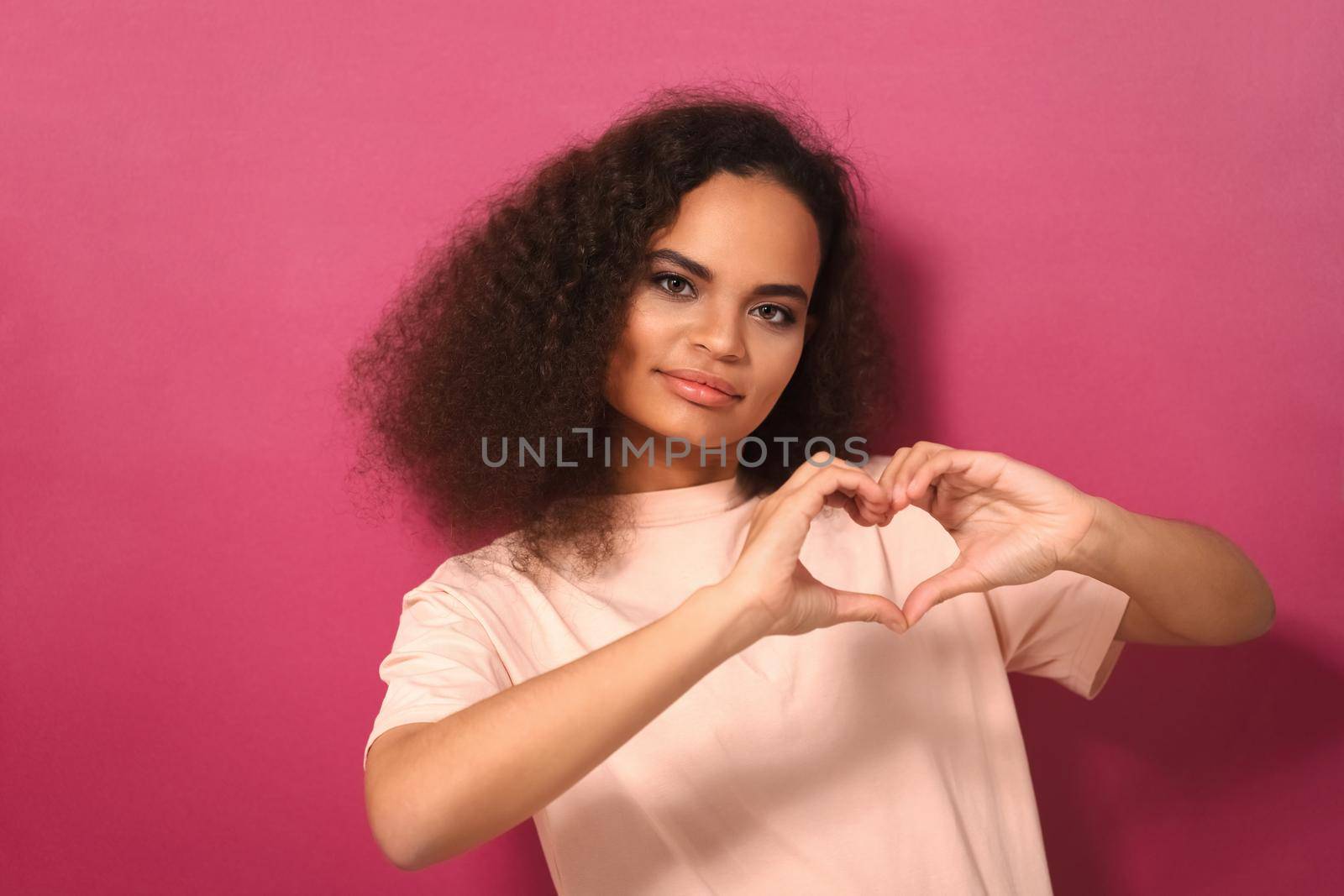 Showing love sign african american girl with long curl hair looking at camera wearing peachy t-shirt isolated on dark pink background. Love concept.