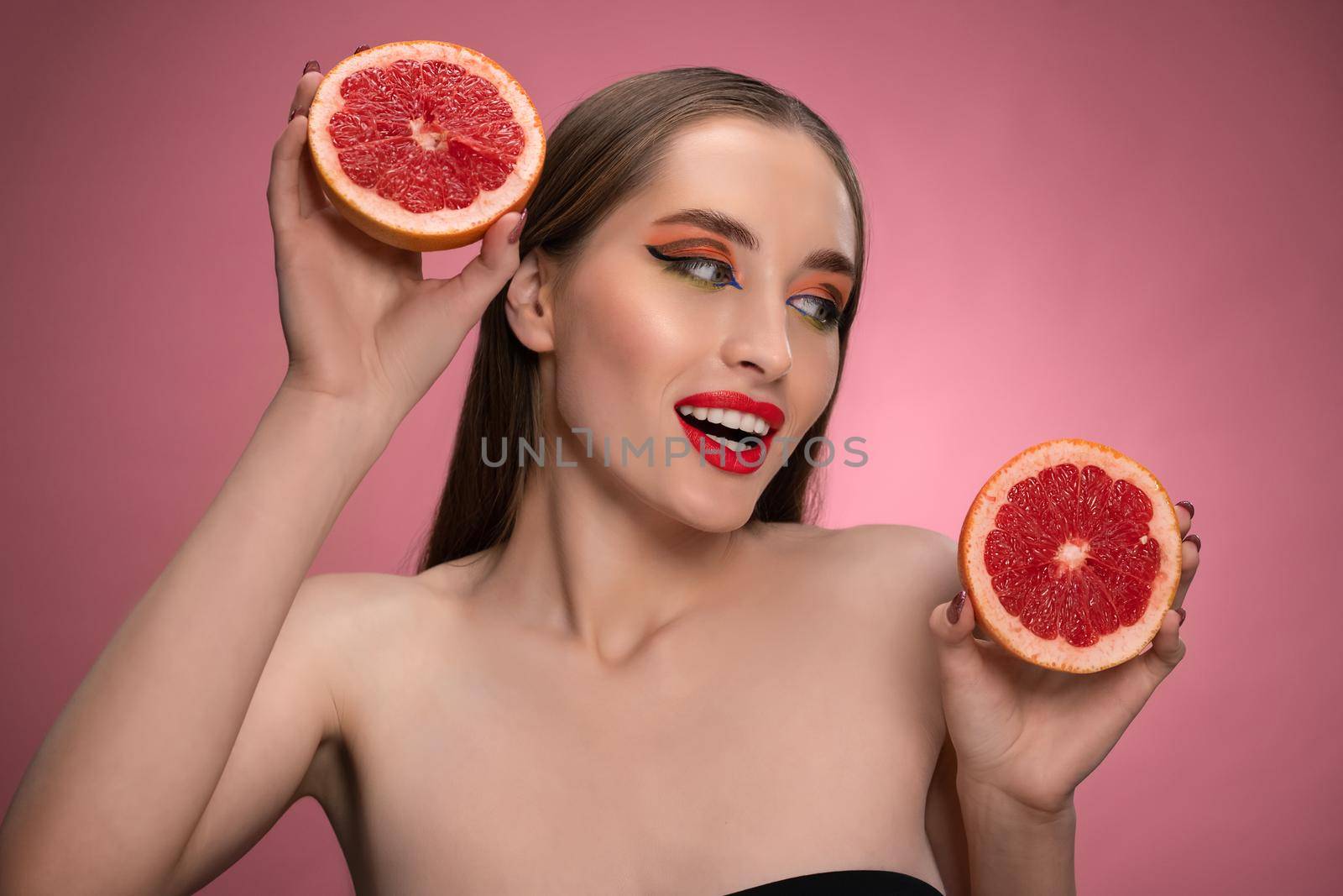 Beautiful young woman model holding two juicy grapefruit sliced in half in her hands looking sideways. Charming joyful funny lady with red lips and long hair isolated on pink background.