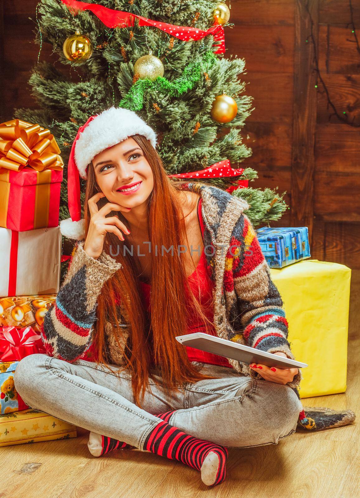 Beautiful Woman in a Winter Knitted Sweater, Hat and Striped Socks Sits on the Floor with Crossed Legs Near a Pile of Gifts and a Christmas Tree. Close-up. High quality photo