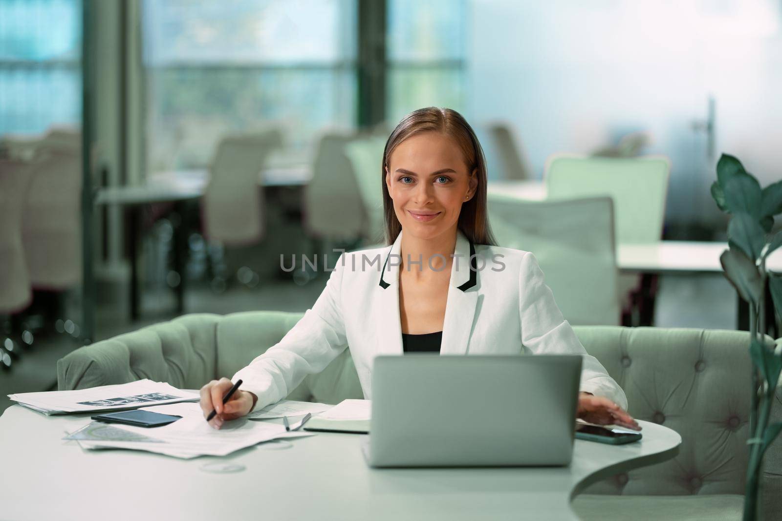 Blond beautiful businesswoman working with documents sitting in front of laptop wearing white official suit. Office worker looking at camera with smile while sitting at her working place.