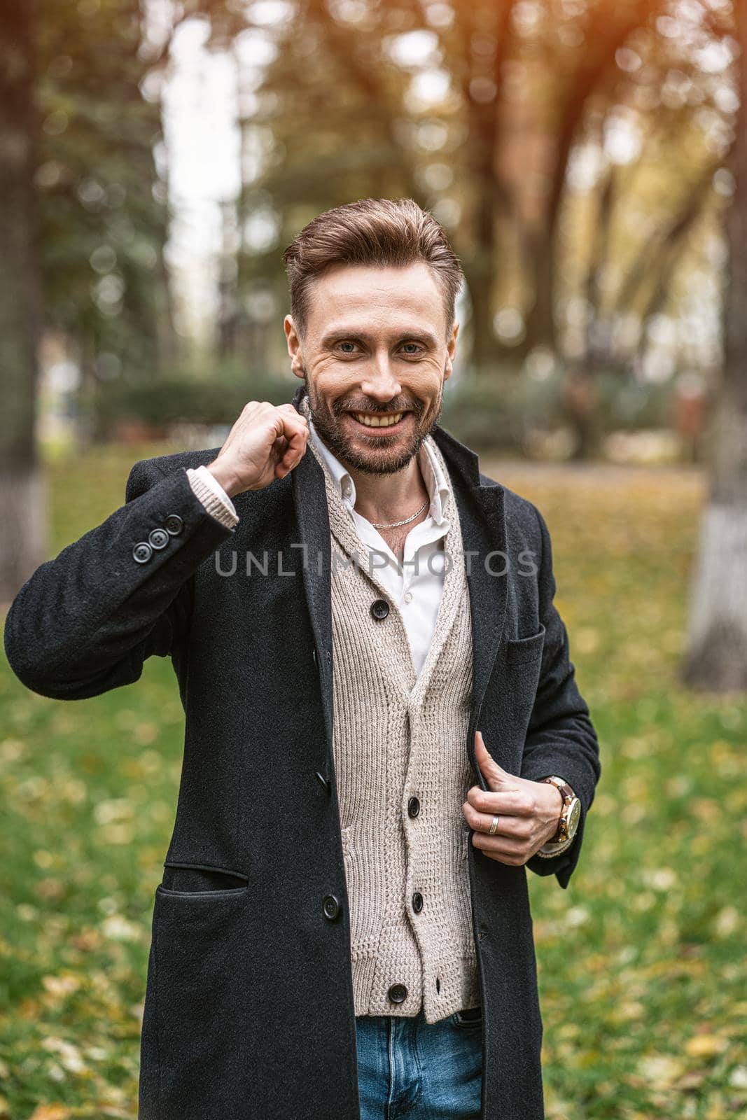Handsome smiling happy man straightens the sides of his dark blue coat. Young freelancer man stands on the street in an autumn coat and cardigan under it looking happy at camera.