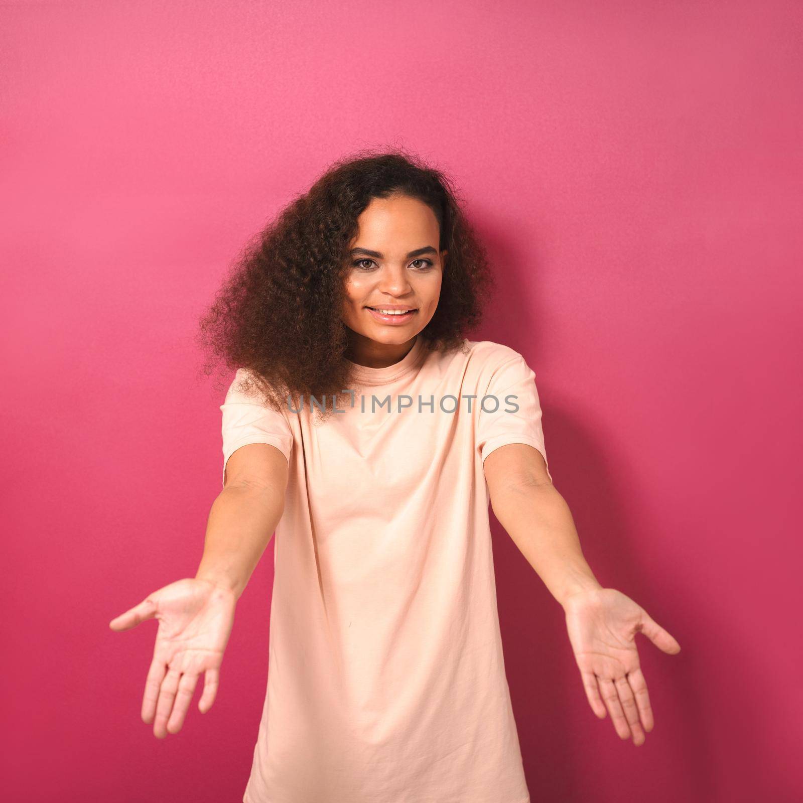 Hands front with palms up a tender looking beautiful young African American woman looking positively at camera wearing peachy t-shirt isolated on pink background. Beauty concept.