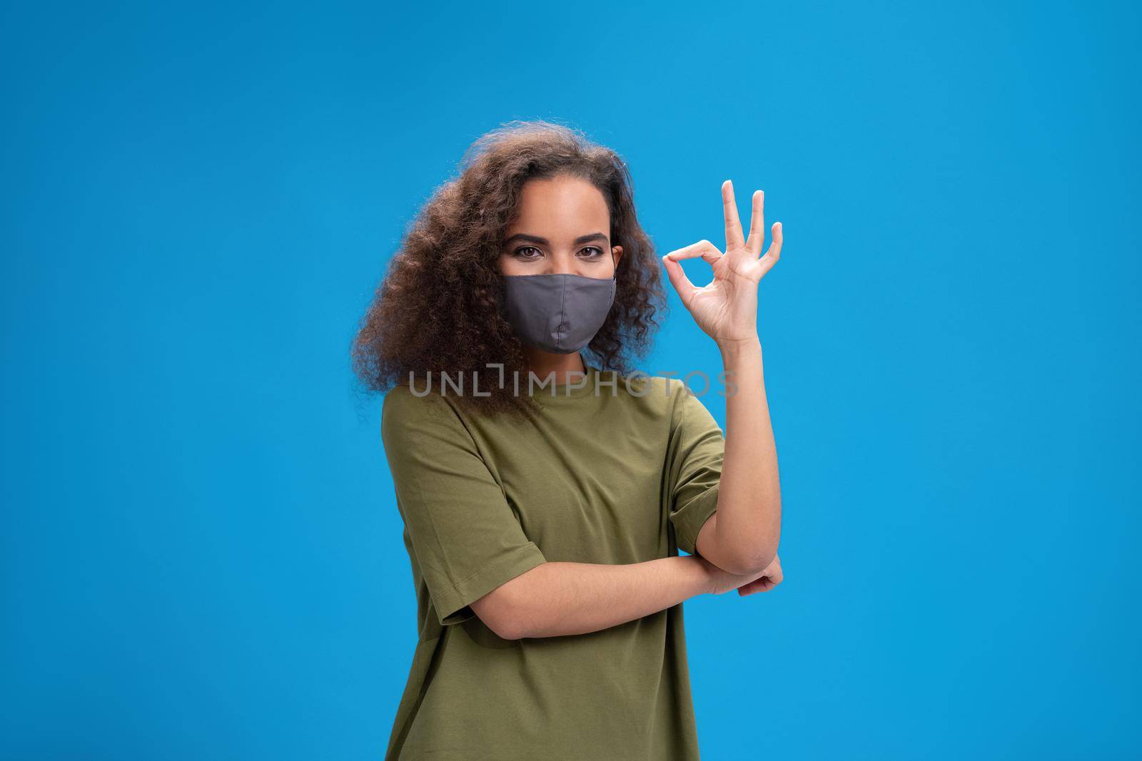 Saying OK using her hands to communicate pretty african american girl in reusable protective face mask. Curly haired young woman showing hello sign talking on camera isolated on blue background.