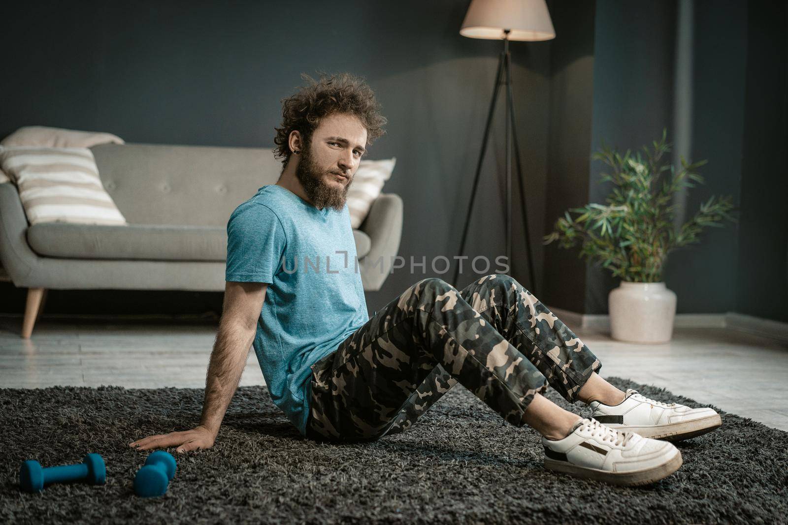Man in a Blue T-shirt Sits on a Carpet Next to his Blue Dumbbells and Gazes at the Camera. Medium Shot. Living Room Background. High quality photo