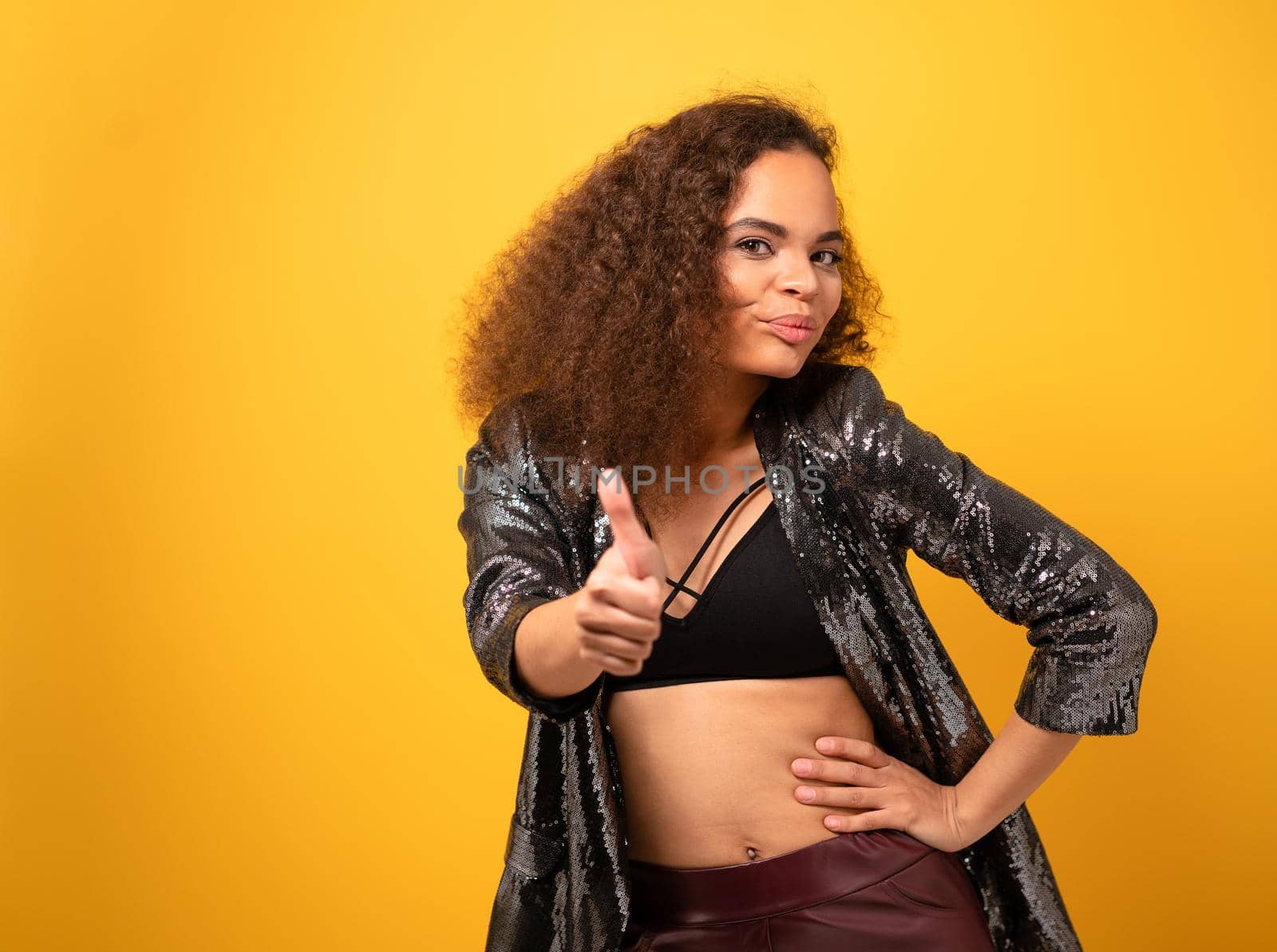 I got you. Beautiful young woman pointing finger with beautiful hairstyle smile looking at camera wearing shiny black jacket and black top. Happy young girl on yellow background.