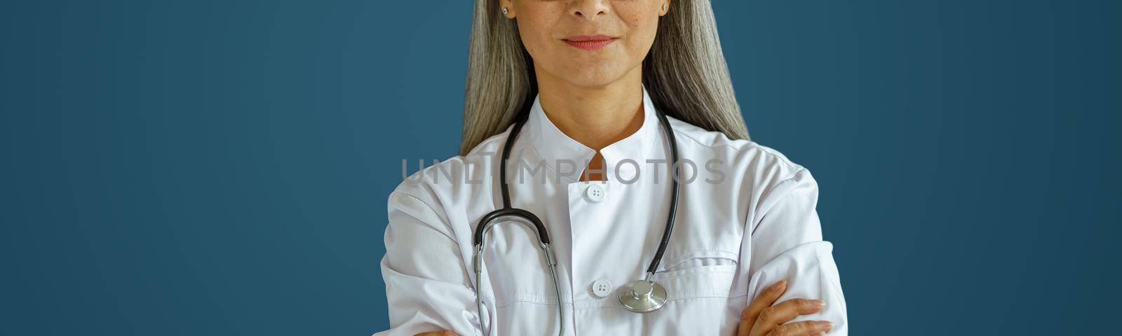Mature female doctor in white robe with glasses and stethoscope stands on blue background in studio. Professional medical staff