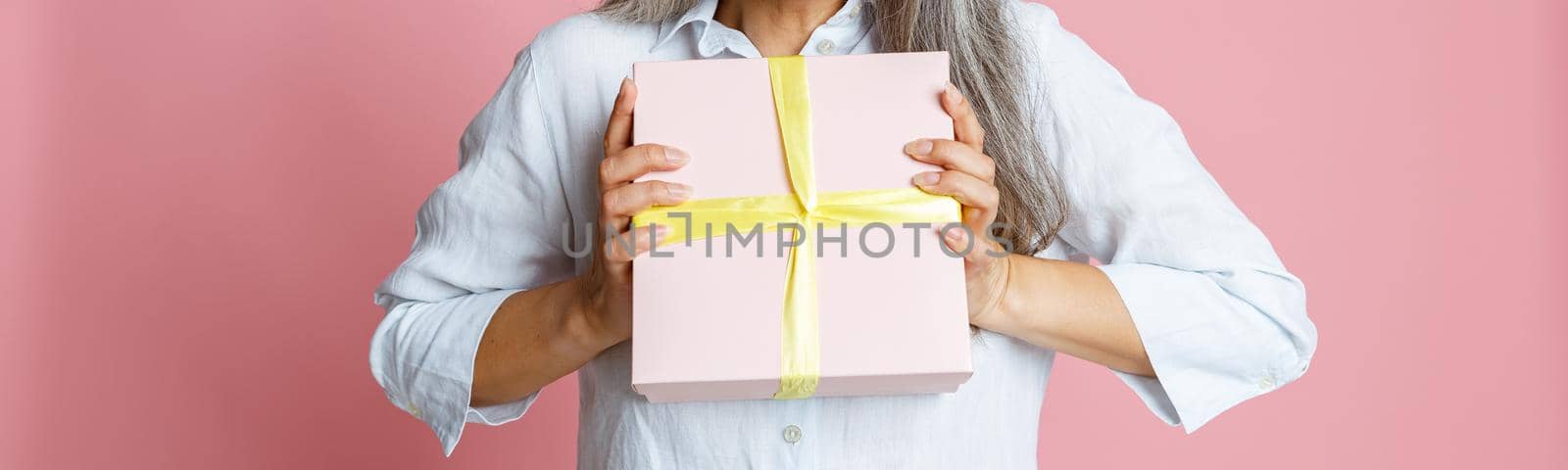 Emotional mature Asian woman with loose grey hair and yellow party hat holds present posing on pink background in studio