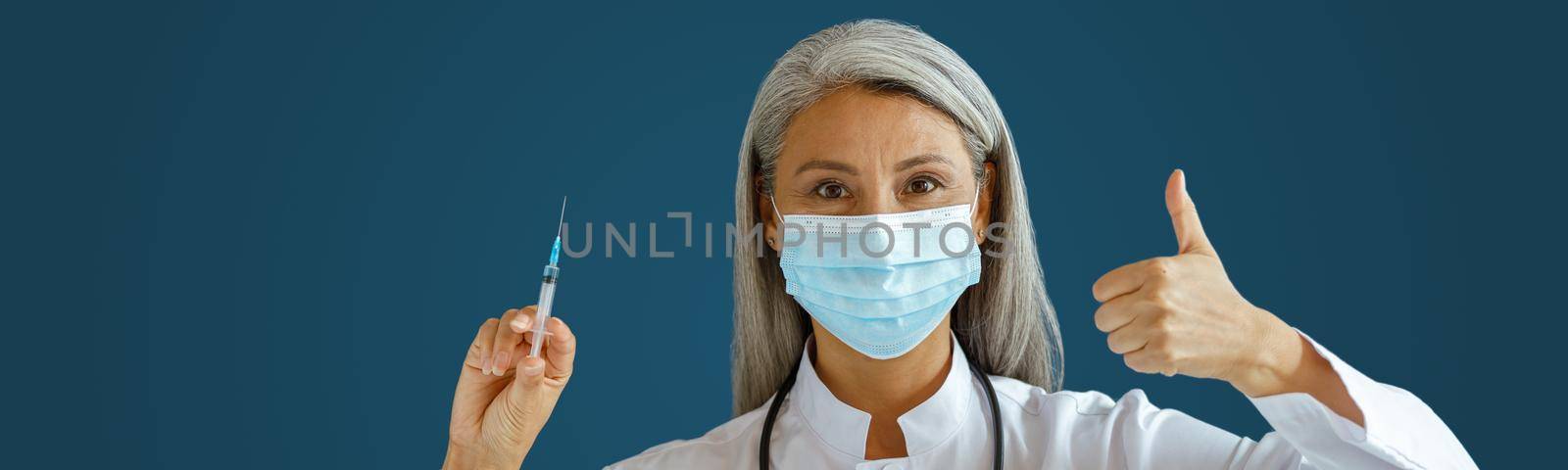 Mature Asian woman doctor in white coat with medical mask holds syringe and shows thumb up standing on blue background in studio. Vaccination day