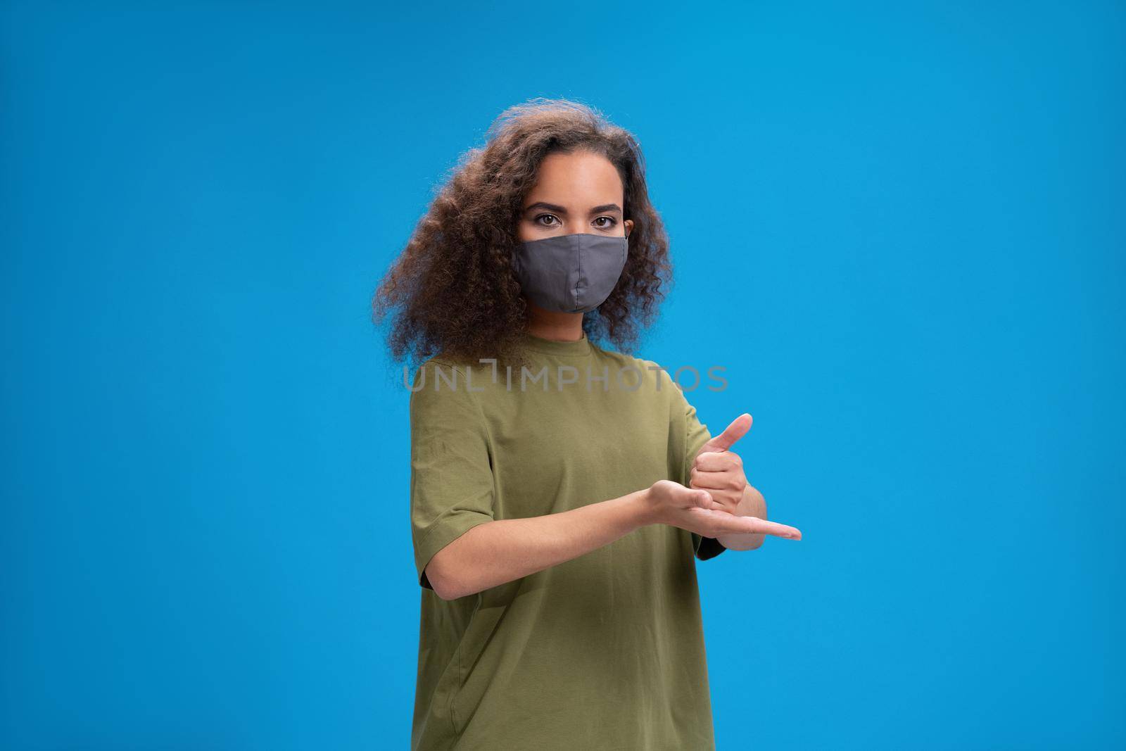 Asking for HELP african american girl using her hands to communicate wearing reusable face protection mask. Curly haired young woman showing hello sign talking on camera isolated on blue background.