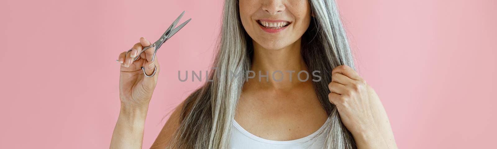 Cheerful Asian lady holds lock of silver hair and scissors on pink background by Yaroslav_astakhov