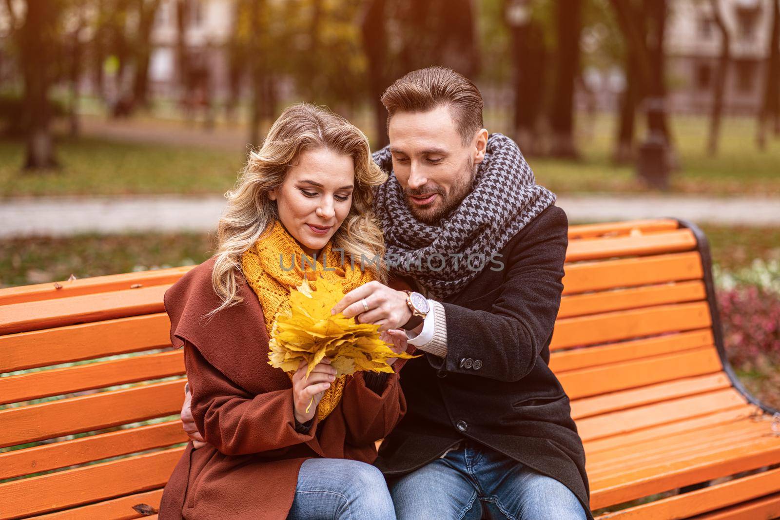 Embracing happy couple sitting gently romantic hugged on a bench in park wearing coats and scarfs Collecting a bouquet of fallen leaves. Love story concept.