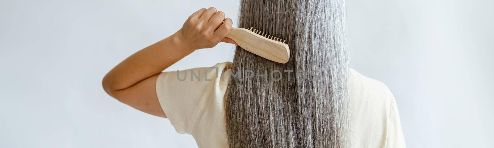 Lady in yellow blouse brushes straight silver hair standing on light grey background in studio backside view. Mature beauty lifestyle