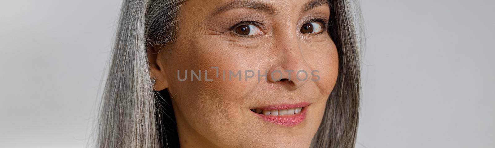 Attractive grey haired lady in stylish shirt smiles to camera on light background in studio. Mature beauty lifestyle