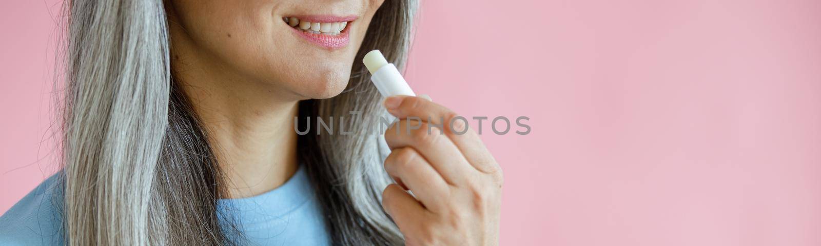 Middle aged grey haired woman in blue t-shirt applies lip balm on pink background in studio closeup, space for text. Mature beauty lifestyle