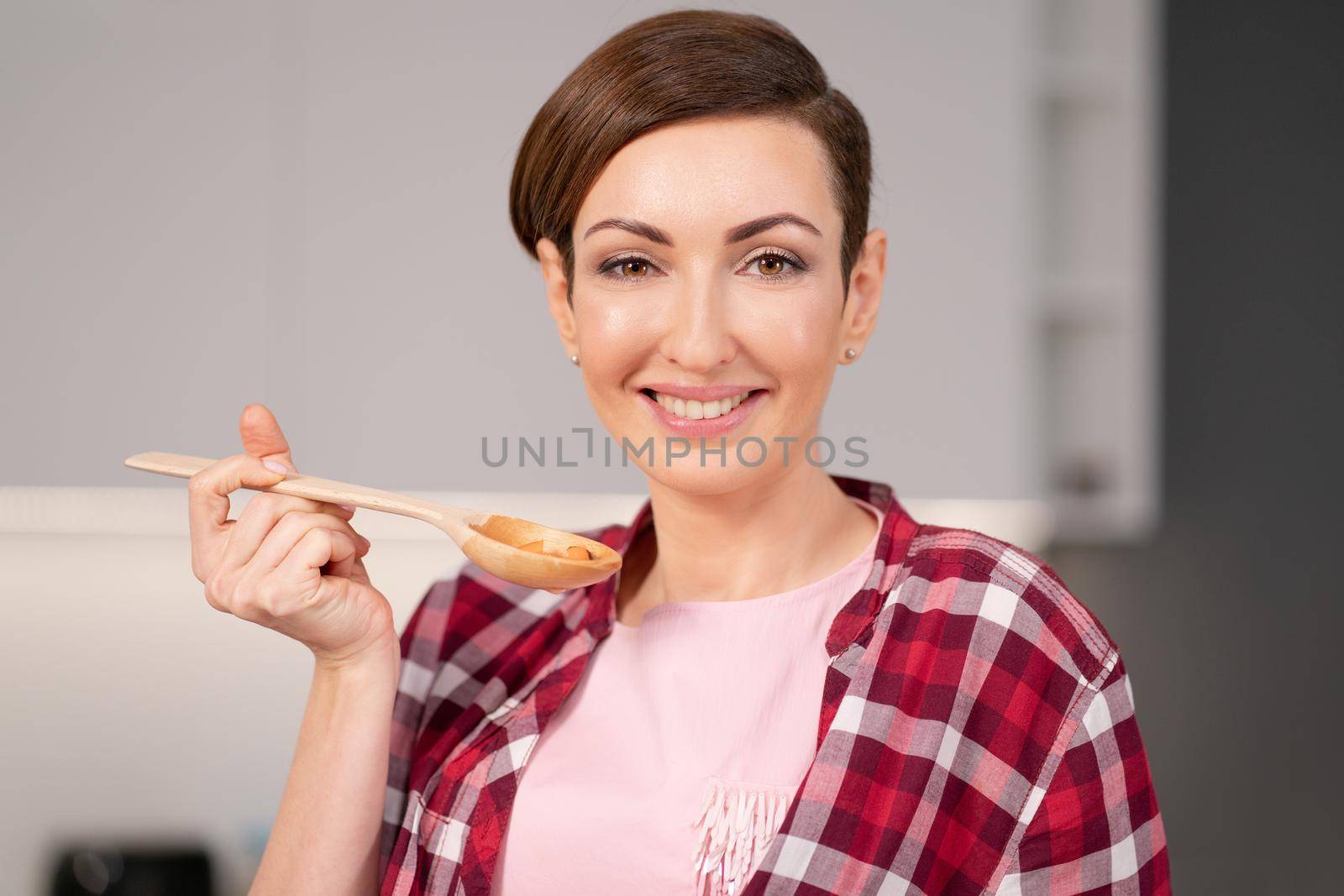 Close up. Charming young housewife with a short hairs cooking a dinner for family trying it using a long wooden spoon standing in the modern kitchen. Healthy food at home.