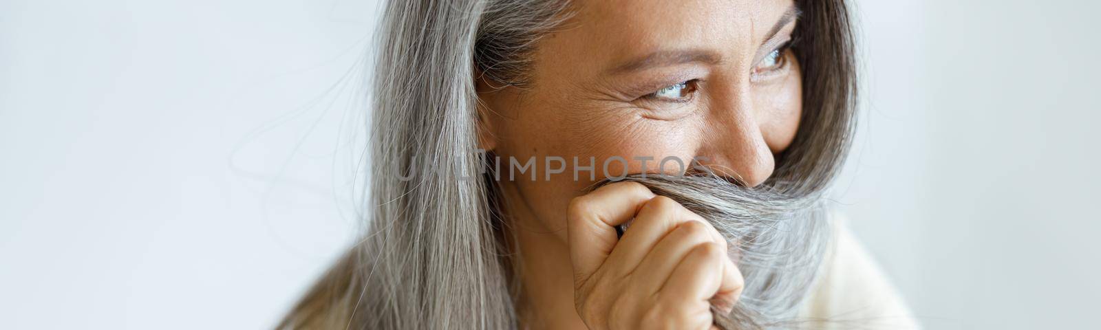 Middle aged Asian woman hides face with grey hair sitting on light background by Yaroslav_astakhov