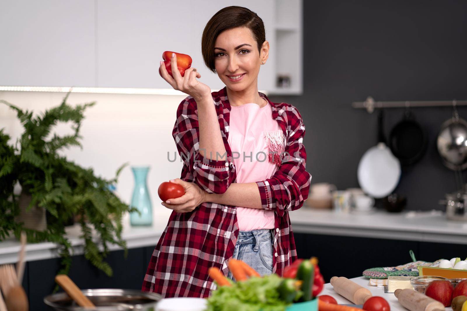 Holding bell pepper and tomato in the hand housewife, wearing plaid shirt with a short hairstyle cooking vegetable ragout at new kitchen in new house. Healthy food at home.