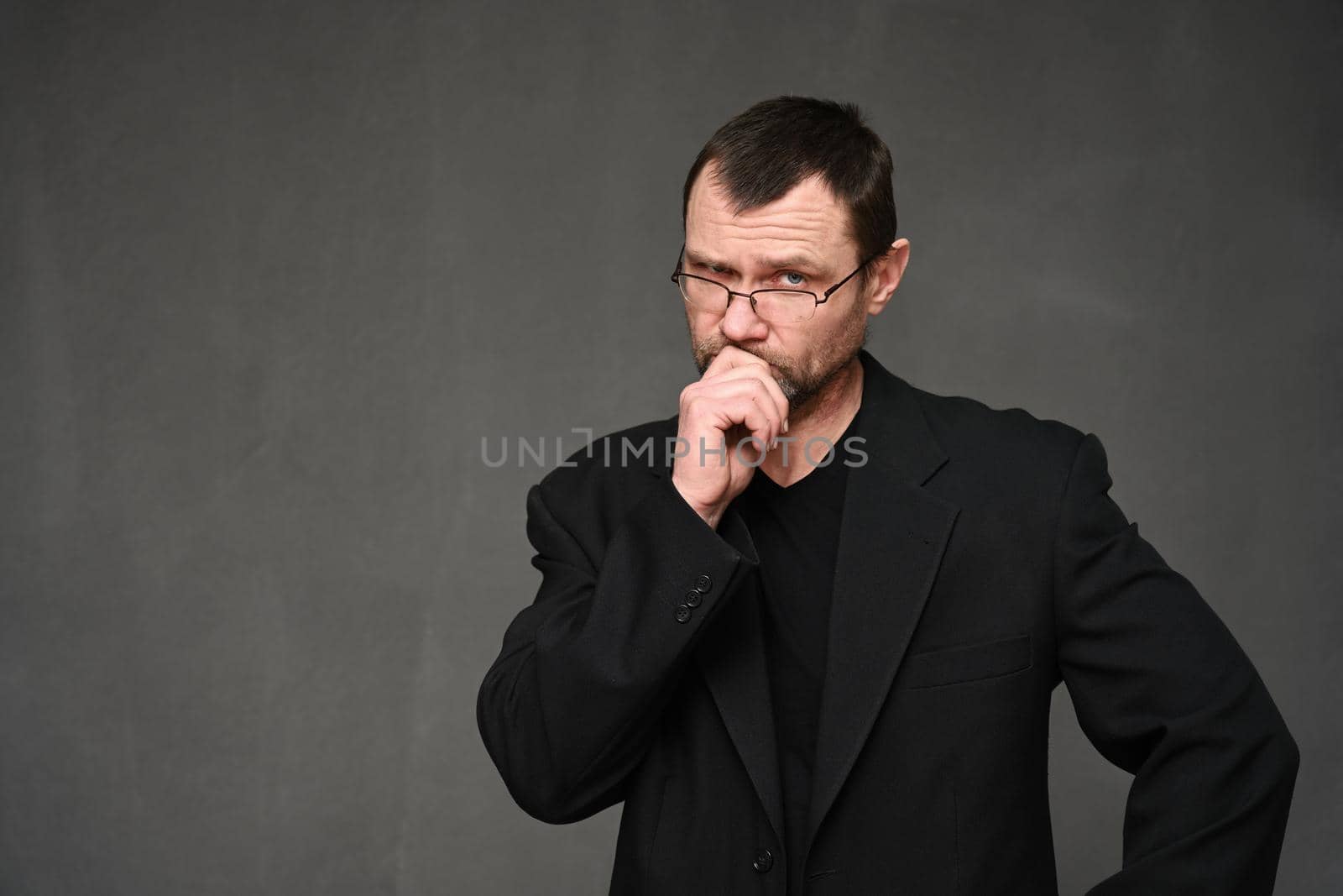 Adult man in a black jacket with glasses looks at the camera thoughtfully on a gray background