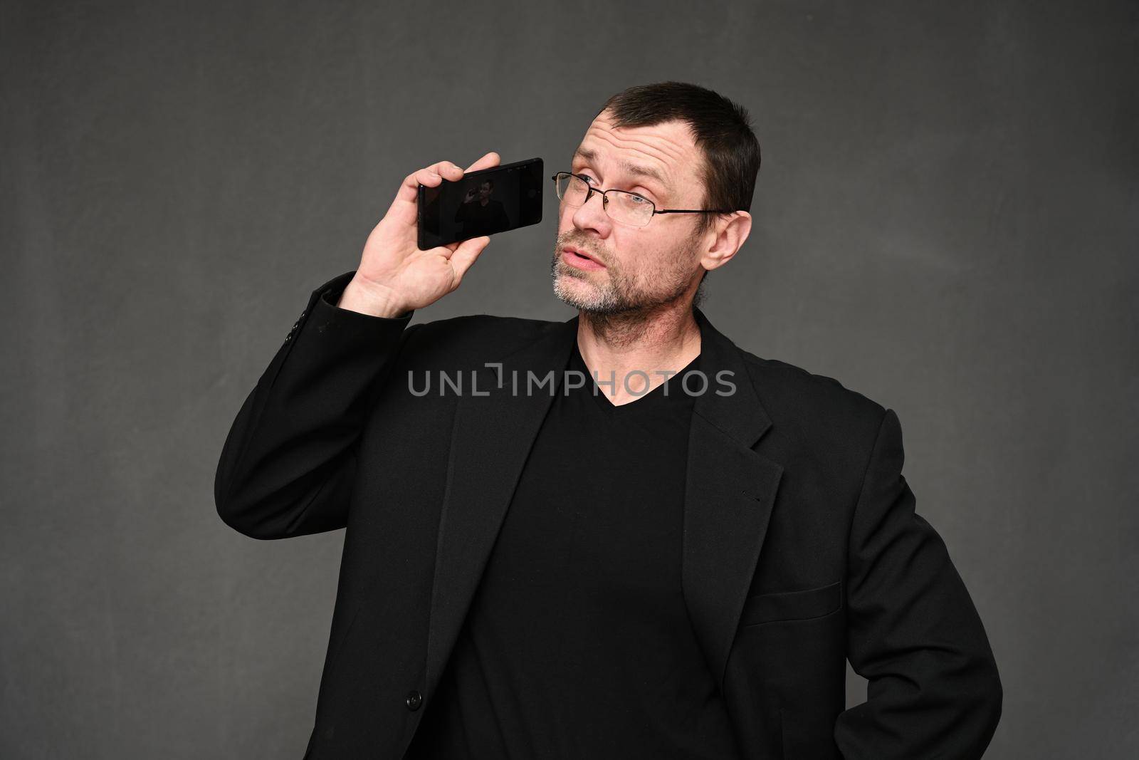 Businessman in a black jacket with glasses talking on the phone with emotions on a gray background