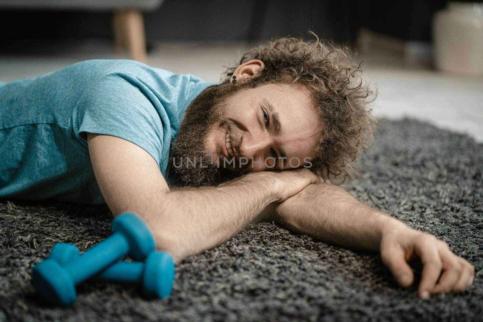 Man Lies Relaxed on the Carpet Next to his Dumbbells. Curly-haired Guy is too Relaxed to Play Sports. Close-up Portrait . High quality photo