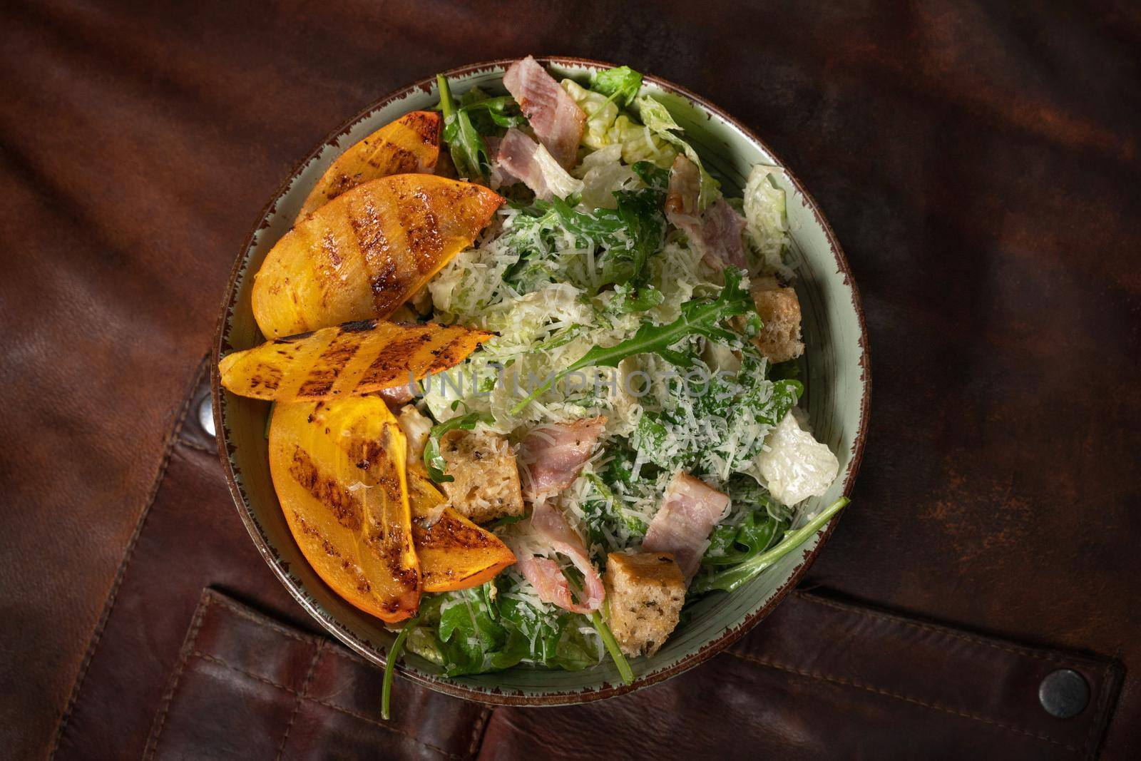 Top view of a bowl of fresh salad with salmon, mozzarella cheese and grilled slices of fruits stands on a table covered with a leather rag. Restaurant concept.