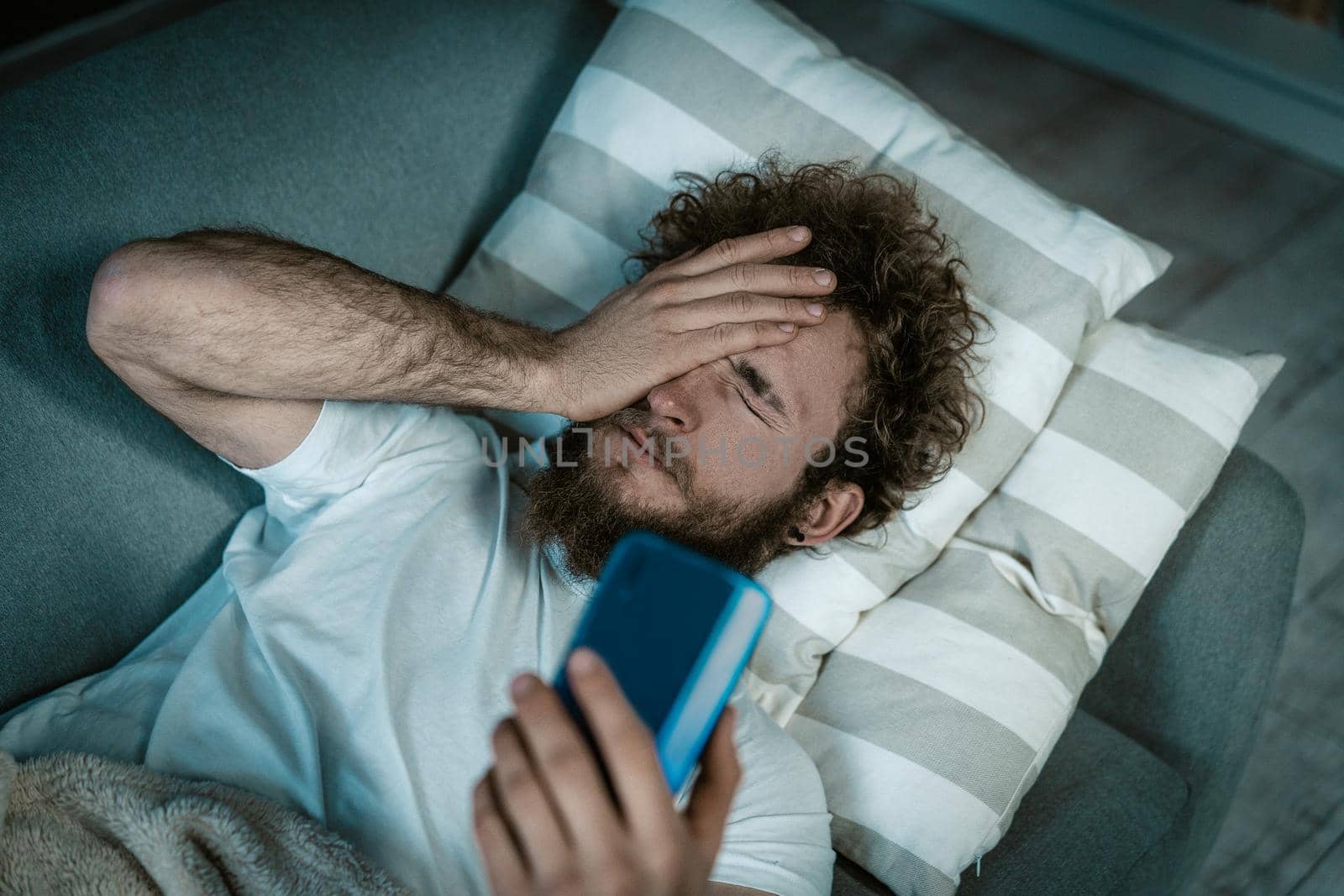 Sleepy Guy Turns Off the Alarm on the Phone. Caucasian Guy Lies on the Bed and Covers Eyes He Has a Smartphone in a Hand. Morning Call, Morning Alarm, Wake up Early. Close-up. Bedroom Background. High quality photo