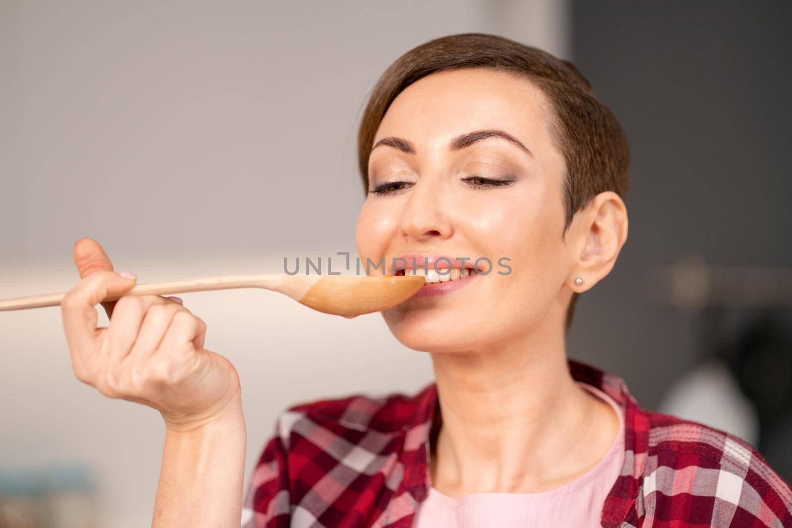 Close-up of a woman trying a cooked dish using a long wooden spoon. Woman with a short hair cooking a dinner for family standing in the modern kitchen of a new house. Healthy food at home.