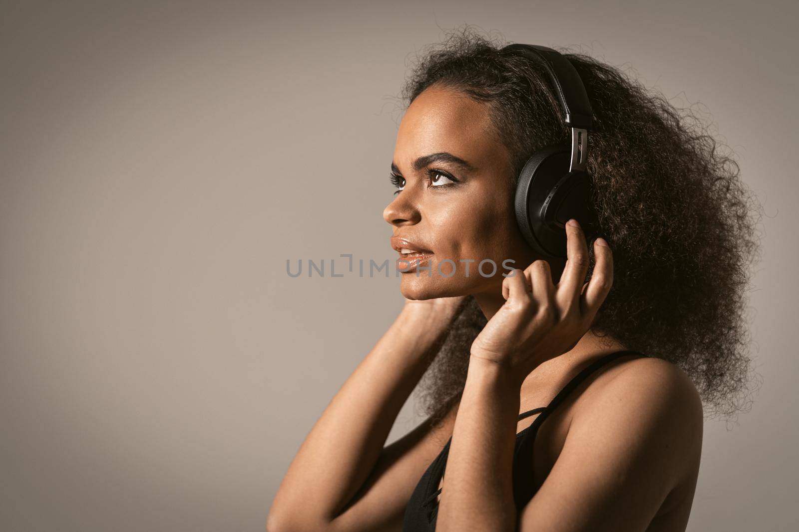 Capture away by music young African-American girl looking up away sideways listening music wearing headphones, isolated on grey background, emotionally move, have fun. Close up.