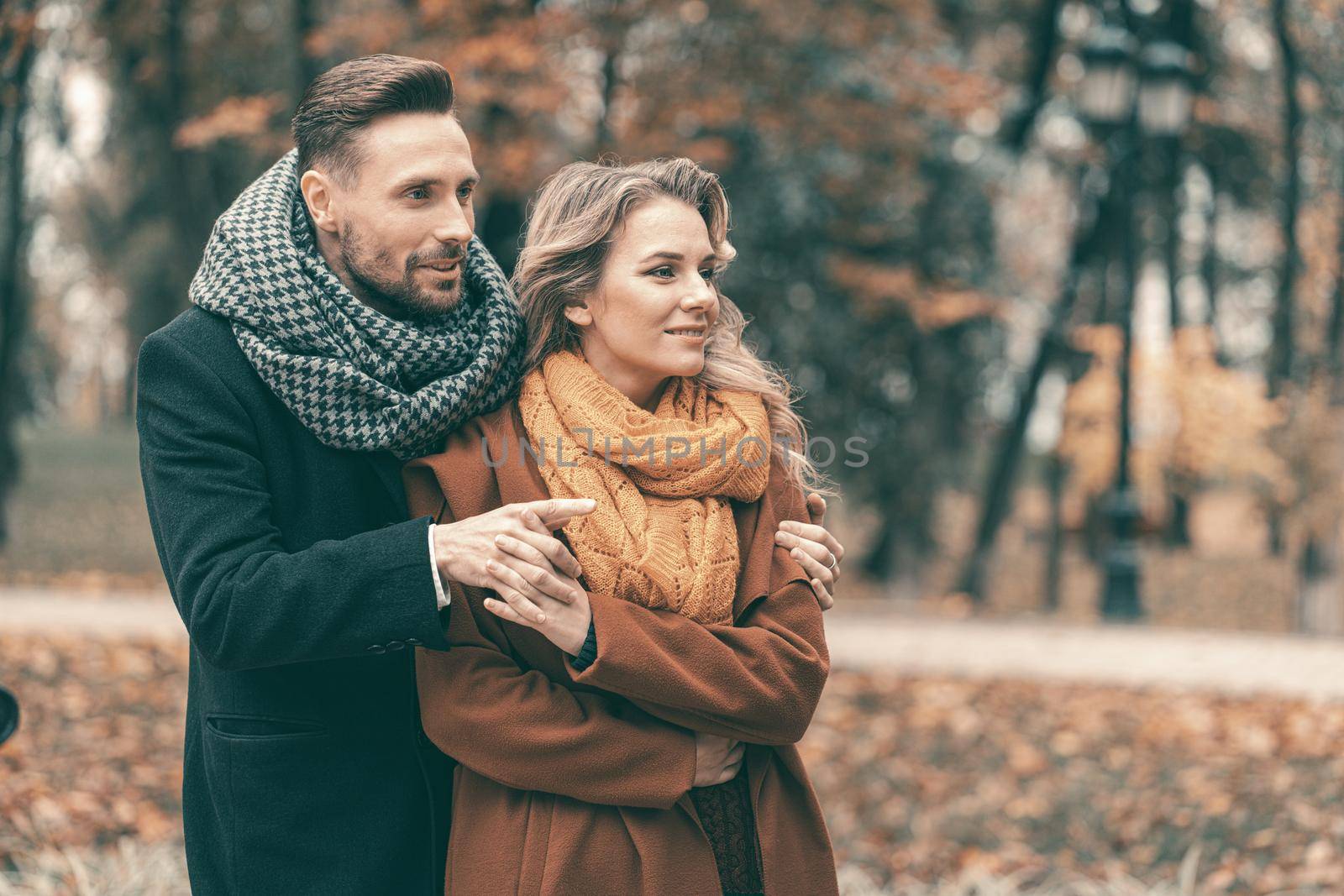 Man hugging woman standing behind her looking one direction sideways in the autumn park. Outdoor shot of a young couple in love having great time having a hug in a autumn park. Close up. Tinted image by LipikStockMedia