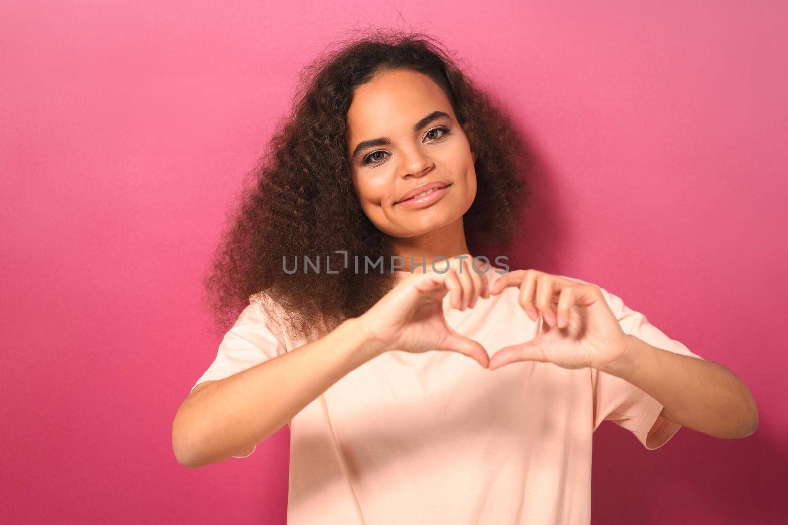Showing heart or love gesture beautiful young African American woman looking positively at camera wearing peachy t-shirt isolated on pink background. Beauty concept.