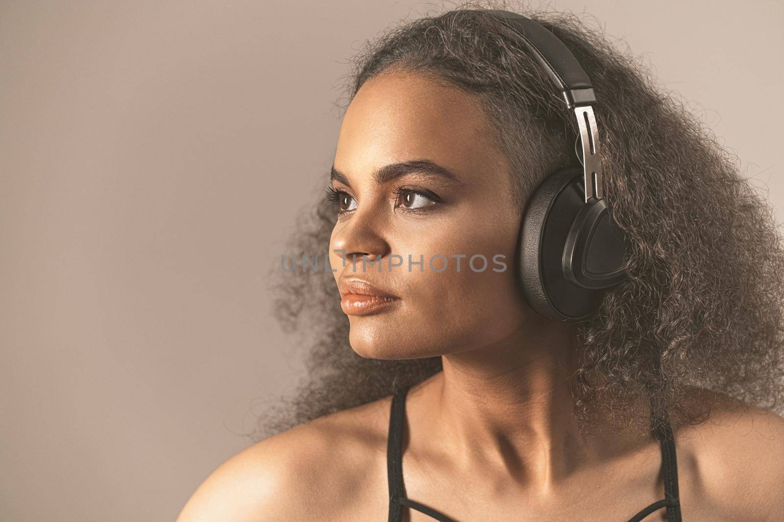 Beautiful young African-American girl listen music looking up away sideways wearing headphones, isolated on beige background. Charming girl with long curl hair listening music. Close up.