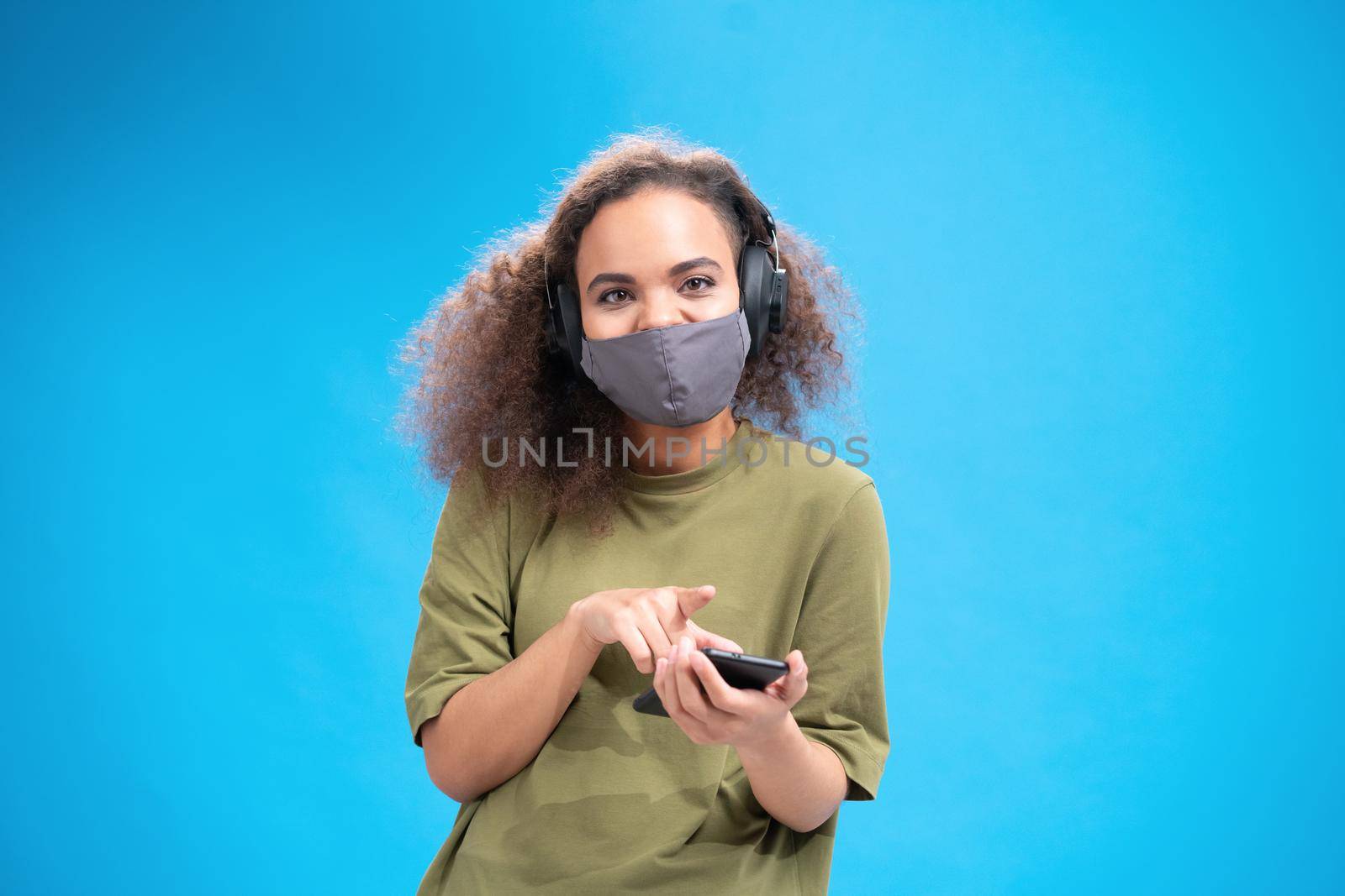 African american girl looking at camera wearing reusable face mask and listen music in headphones. Wearing safety mask while using smartphone to communicate on social distancing isolated on blue.