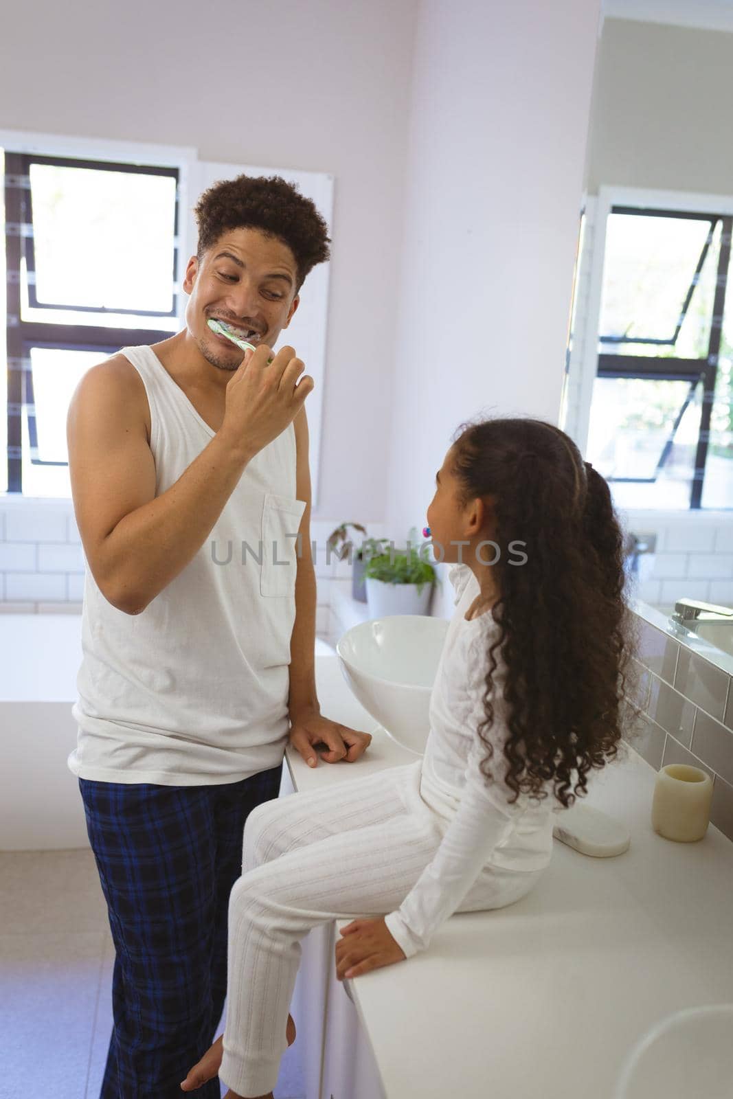 Cheerful biracial man brushing teeth while looking at daughter sitting on bathroom counter by Wavebreakmedia