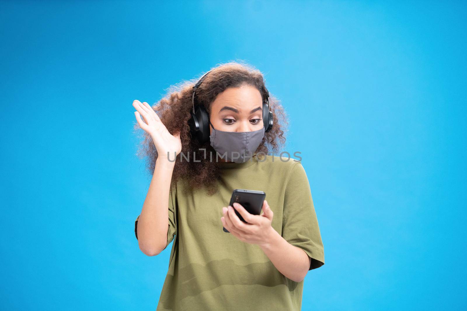 Gesturing african american girl looking at camera wearing reusable face mask and headphones. Wearing safety mask while using smartphone to communicate on social distancing isolated on blue.