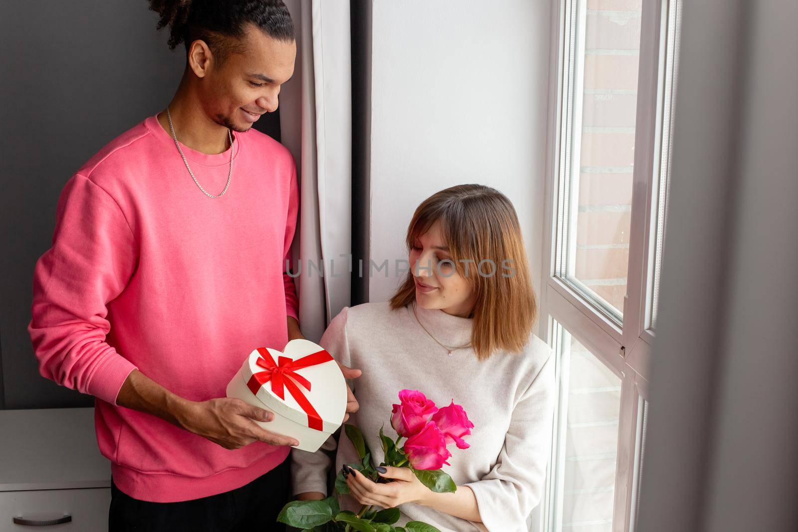 A stylish guy gives his girlfriend, a white heart-shaped box with a bow and a bouquet of roses by Zakharova