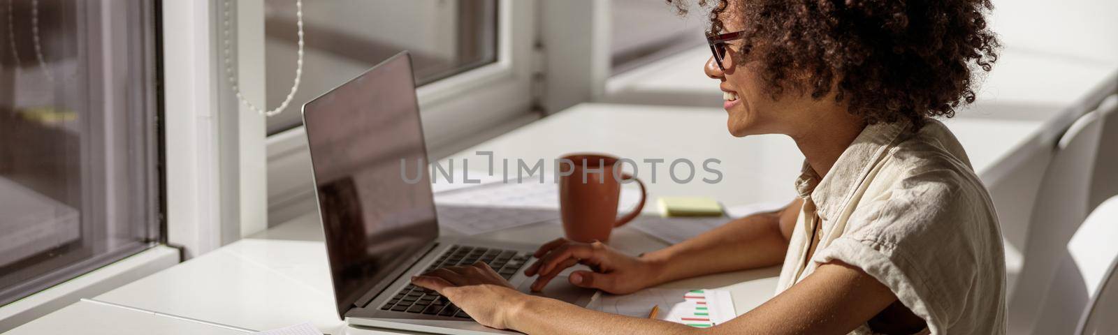 Smiling concentrated female administrative typing messages on laptop while sitting near the window in the office