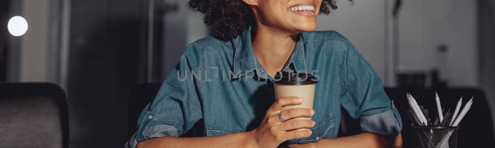 Smiling young multiethnic lady sitting at work desk and holding cup of coffee in the office