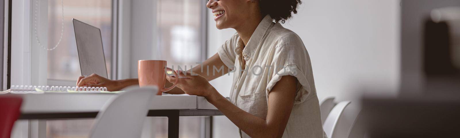 Side view of happy African American woman holding stickers and working with laptop near the window