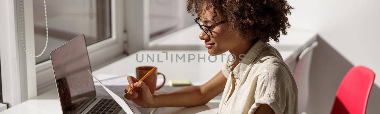 Happy pretty woman wearing glasses and sitting at workplace while holding document and reading