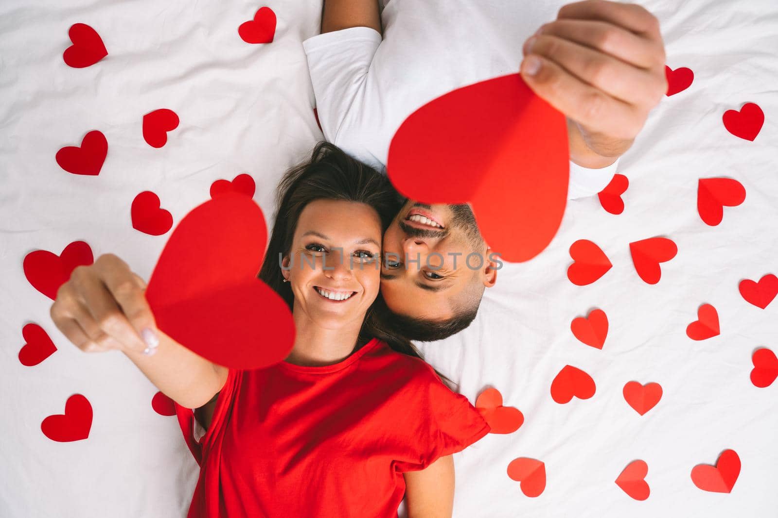 Top view of happy young couple in love holding red paper hearts while lying on the white bed at home. High quality photo