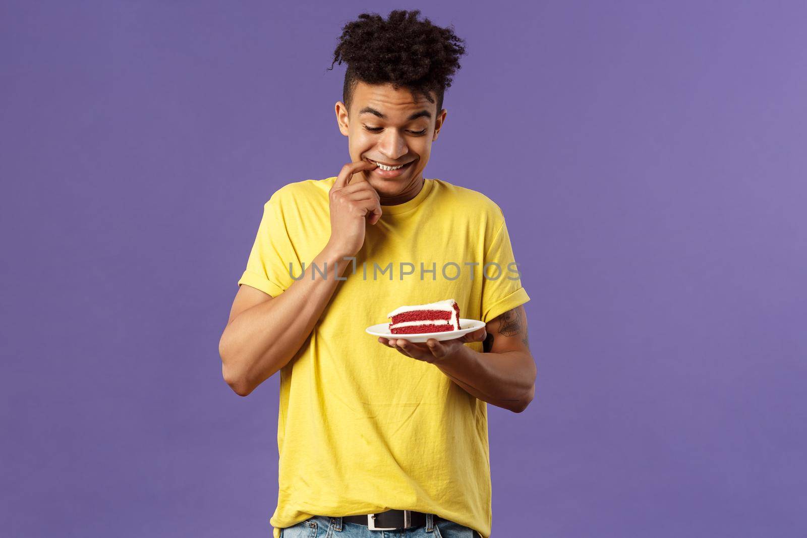 Celebration, party and holidays concept. Portrait of boyfriend cant resist temptation to eat last piece of cake, biting lips and smiling eager to have bite of dessert, hesitating, purple background by Benzoix