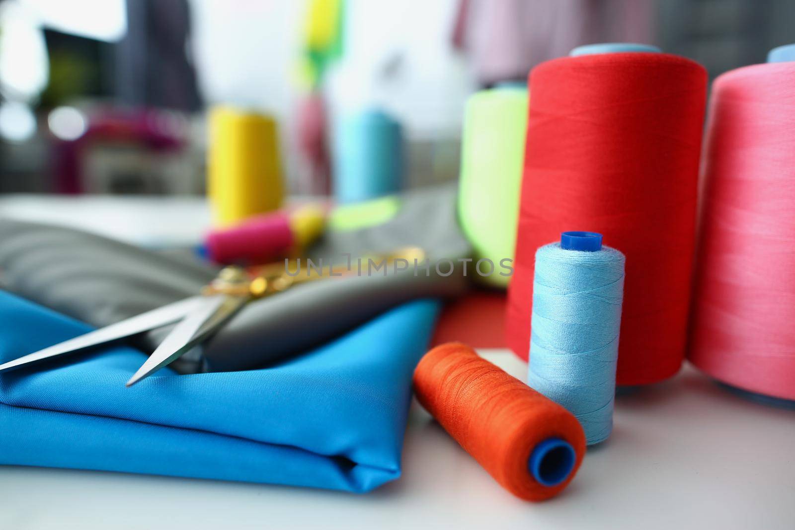 Close-up of bright sewing accessories, scissors and threads on blue cotton fabric, creative working space in private atelier. Hobby, art, sewing concept