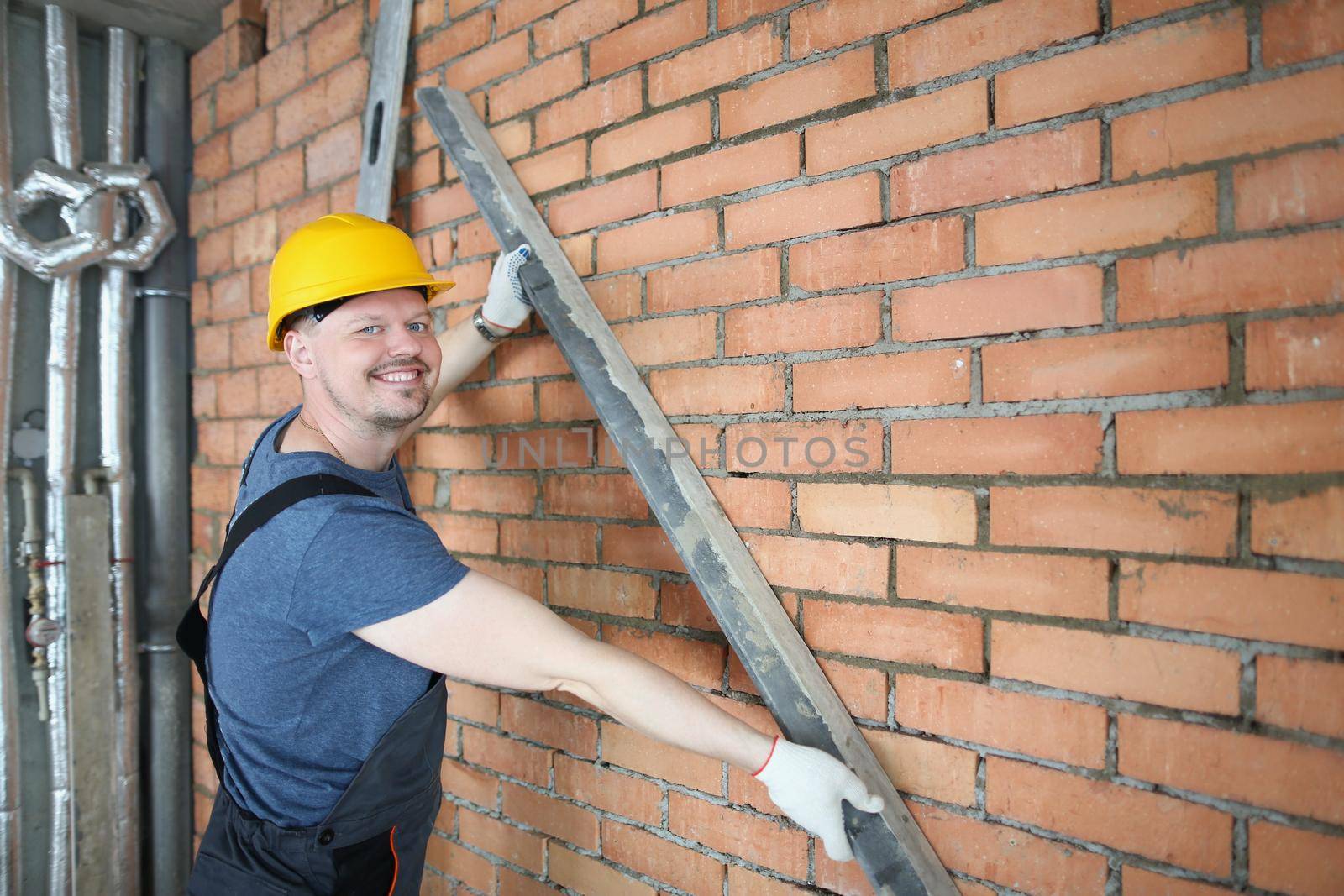 Portrait of happy constructor hold metal detail close to brick wall, concrete red wall in empty room. Creating house, building process. Renovation concept