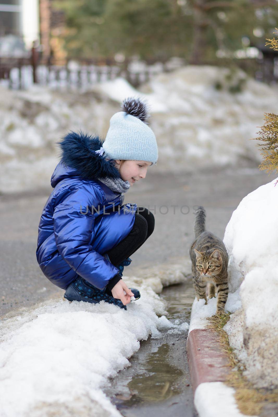 Little girl in blue coat playing with cat on melting snow, puddles and ice. Spring thaw in early spring