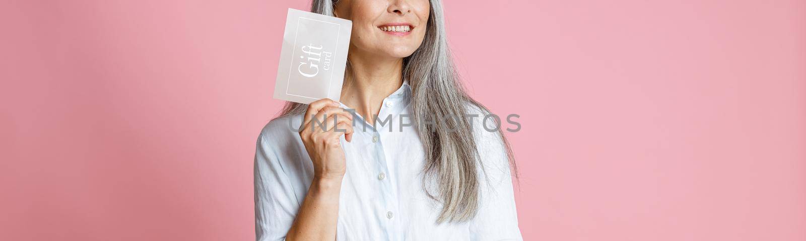 Pretty cheerful silver haired Asian woman holds gift card having ideas on pink background in studio. Shopping certificate