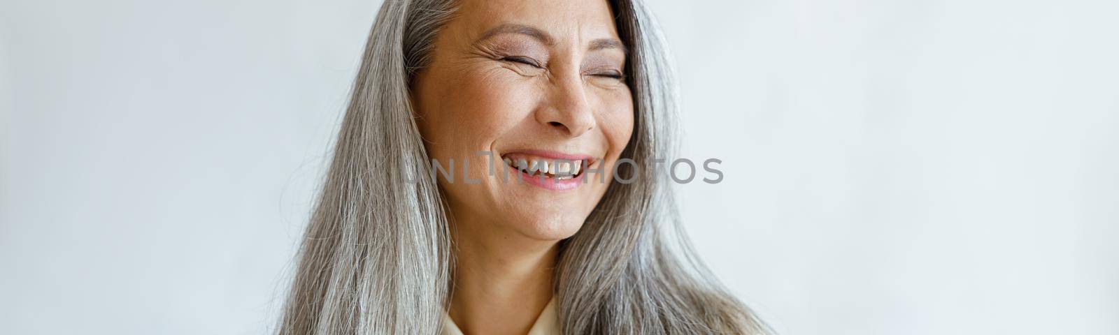 Pretty laughing Asian lady with natural silver hair poses for camera on light grey background in studio. Mature beauty lifestyle