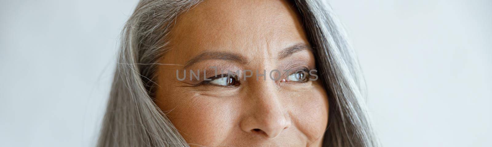 Attractive middle aged Asian lady with loose grey hair poses for camera on light background in studio closeup. Mature beauty lifestyle