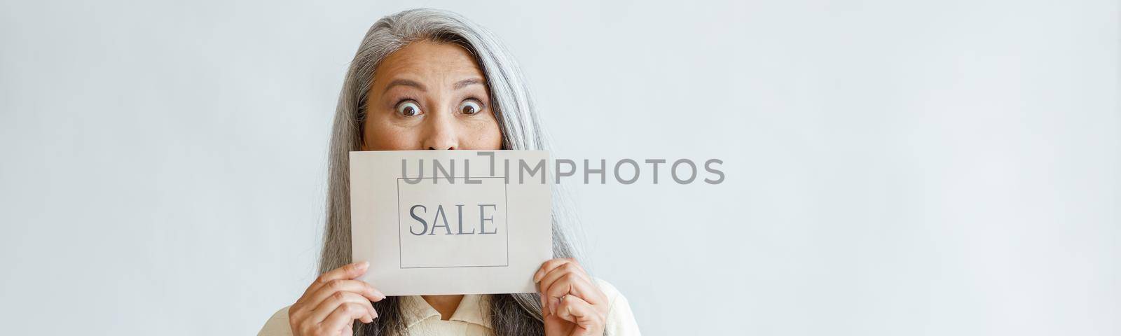 Shocked silver haired Asian lady holds card with word Sale on light grey background in studio, space for text