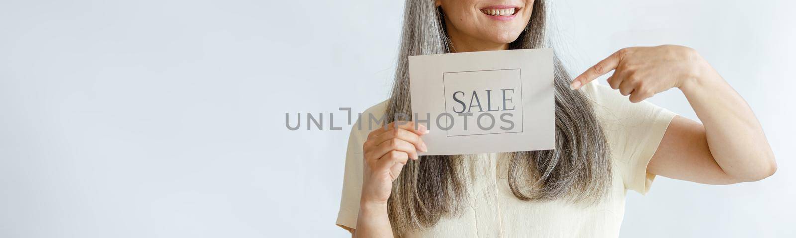 Joyful middle aged Asian woman with long grey hair points onto Sale sign posing on light background in studio, space for text