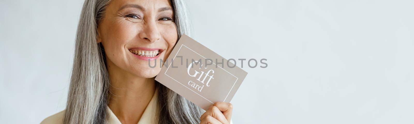 Pretty smiling mature Asian lady holds gift card posing on light grey background in studio, space for text. Shopping certificate